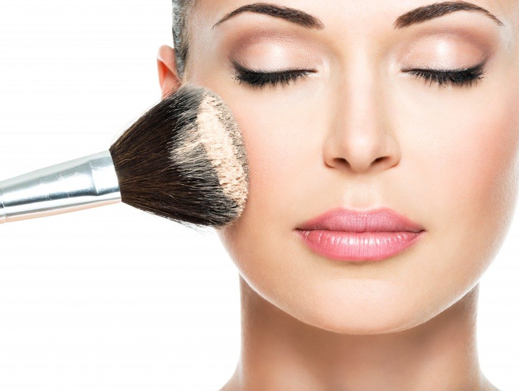 Closeup portrait of a woman applying dry cosmetic tonal foundation on the face using makeup brush
